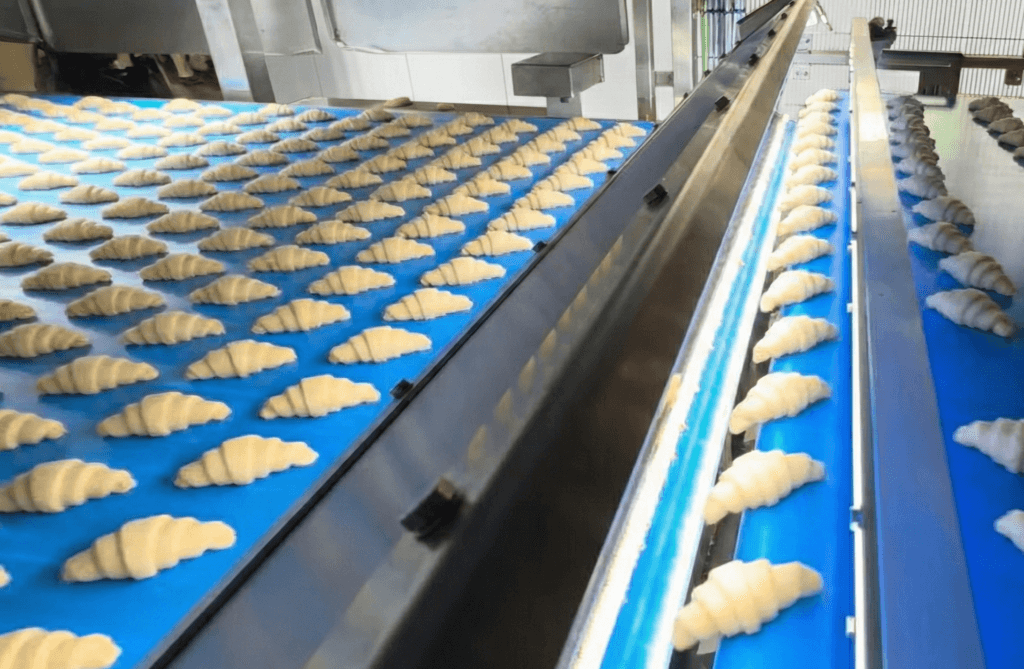 Rows of freshly formed croissants on a blue conveyor belt being analyzed by photonic quality control systems.
